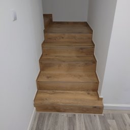 EXCELLENT Michał Mączka - Wykończone schody z drewnopodobnych paneli, kontrastujące z szarą podłogą imitującą drewno. Białe ściany i listwy przypodłogowe. Widok z góry na dół.