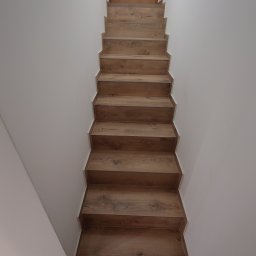 EXCELLENT Michał Mączka - Nowoczesne schody drewniane z wyraźnym rysunkiem słojów, prowadzące do drewnianych drzwi z małym, romboidalnym okienkiem, na tle białych ścian.