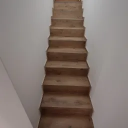 Nowoczesne schody drewniane z wyraźnym rysunkiem słojów, prowadzące do drewnianych drzwi z małym, romboidalnym okienkiem, na tle białych ścian.