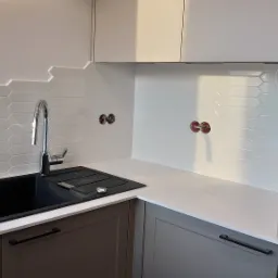 Nowoczesna kuchnia z czarnym zlewozmywakiem, chromowaną baterią, białymi heksagonalnymi płytkami i szarymi szafkami z czarnymi uchwytami, widoczne gniazdka elektryczne przed montażem.