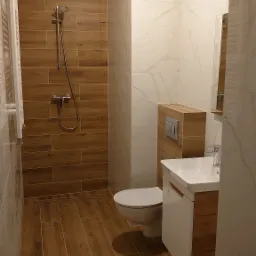 Wykończona łazienka z brązowymi i białymi płytkami, kabiną prysznicową z deszczownicą, podwieszaną toaletą i umywalką z szafką w odcieniach drewna.