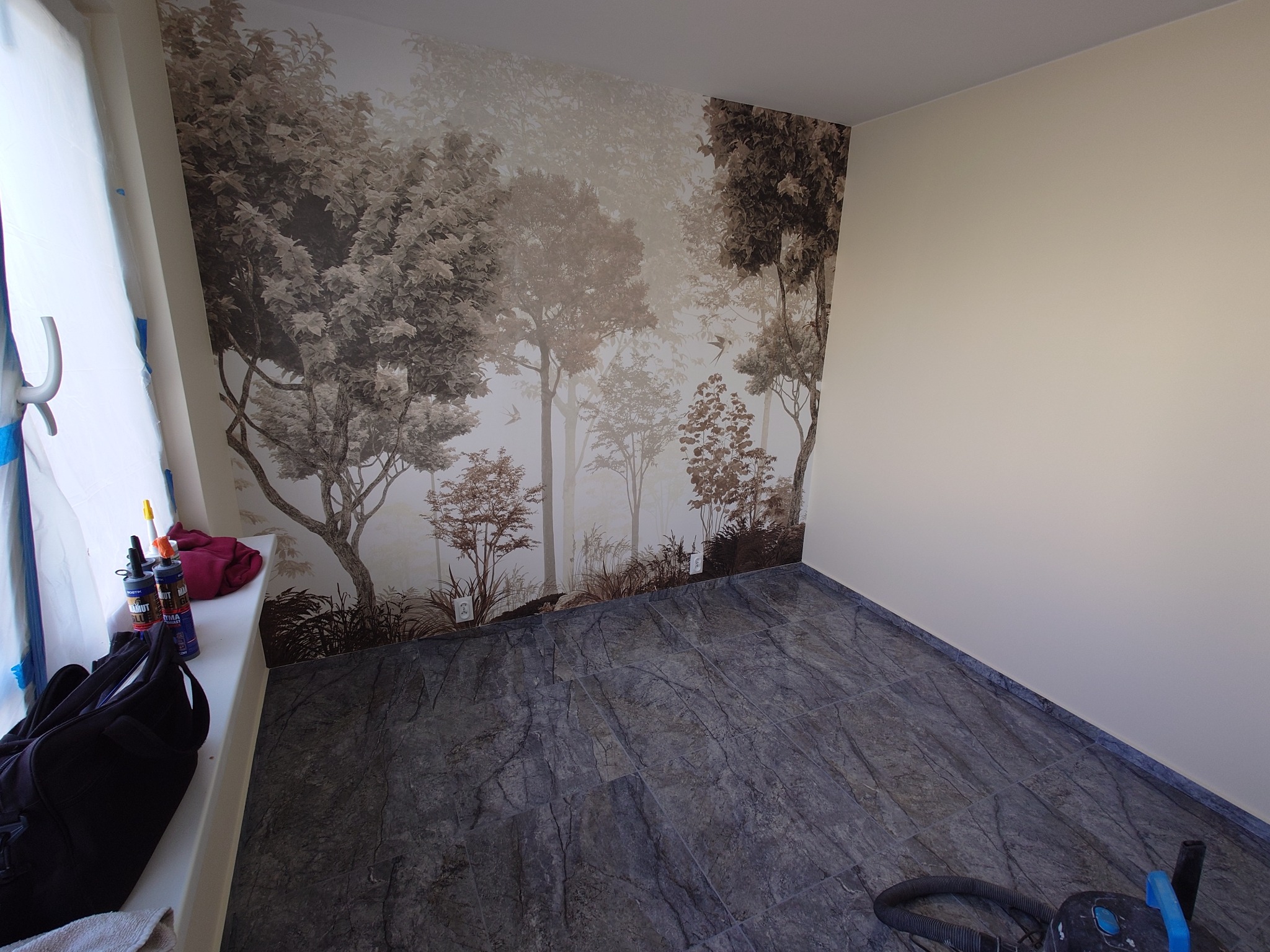 Pokój po tapetowaniu z fototapetą leśną i ułożonymi szarymi płytkami imitującymi kamień. Widoczny odkurzacz i akcesoria malarskie.