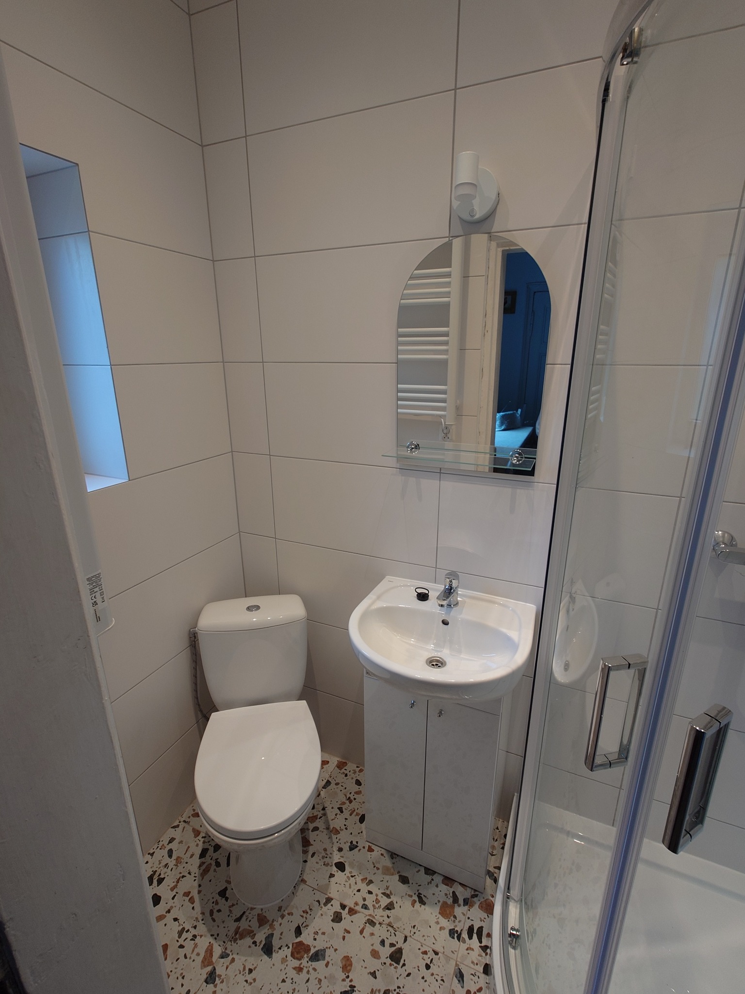 Jasna, wykafelkowana łazienka z toaletą, umywalką z szafką i kabiną prysznicową. Na podłodze wzorzysta mozaika. W lustrze odbicie grzejnika.