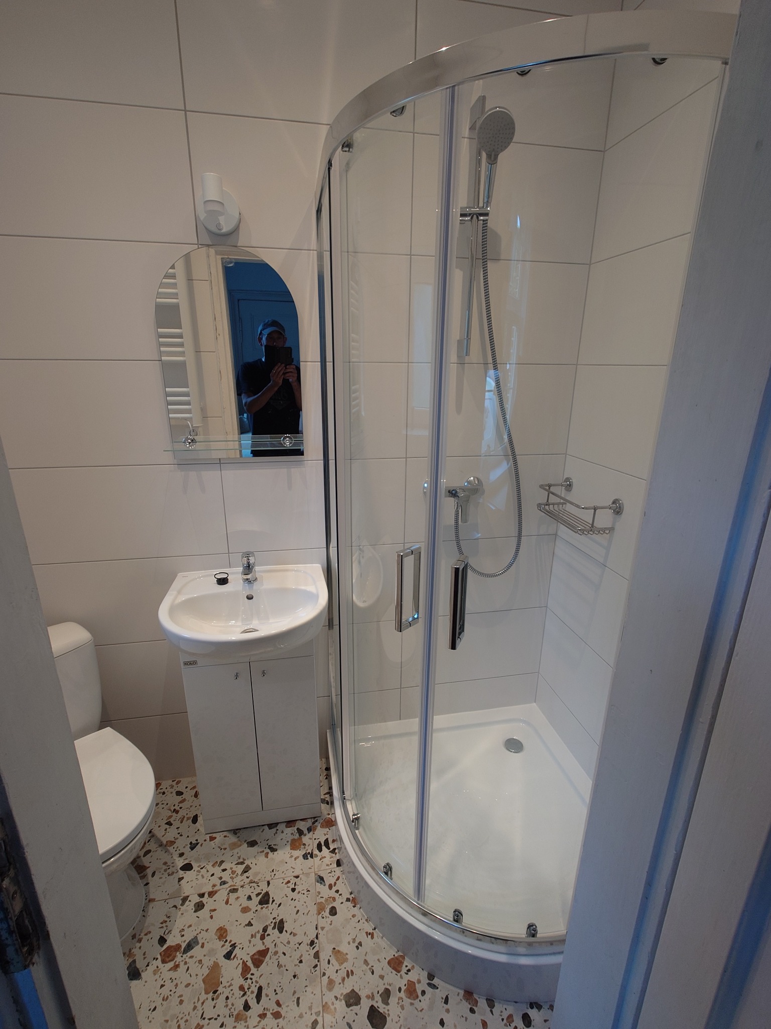 Nowoczesna łazienka z kabiną prysznicową, umywalką z szafką i lustrem. Białe płytki na ścianach, mozaika na podłodze. W lustrze odbija się osoba robiąca zdjęcie.