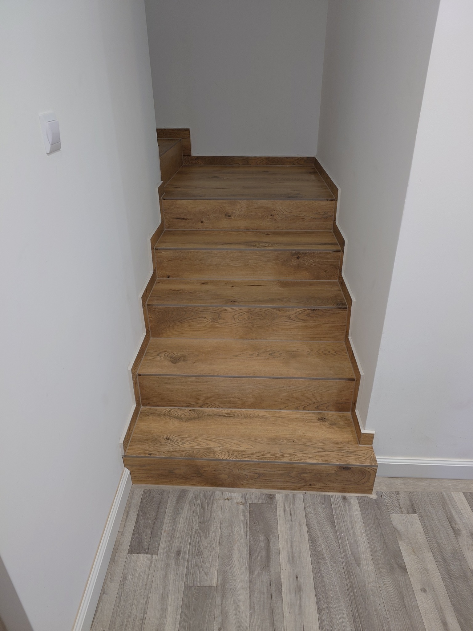 Wykończone schody z drewnopodobnych paneli, kontrastujące z szarą podłogą imitującą drewno. Białe ściany i listwy przypodłogowe. Widok z góry na dół.