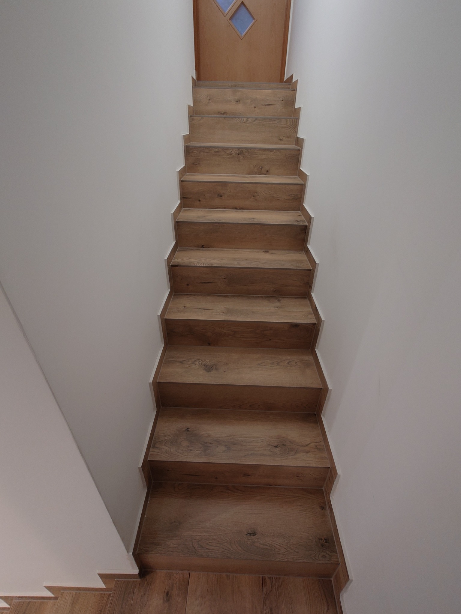 Nowoczesne schody drewniane z wyraźnym rysunkiem słojów, prowadzące do drewnianych drzwi z małym, romboidalnym okienkiem, na tle białych ścian.