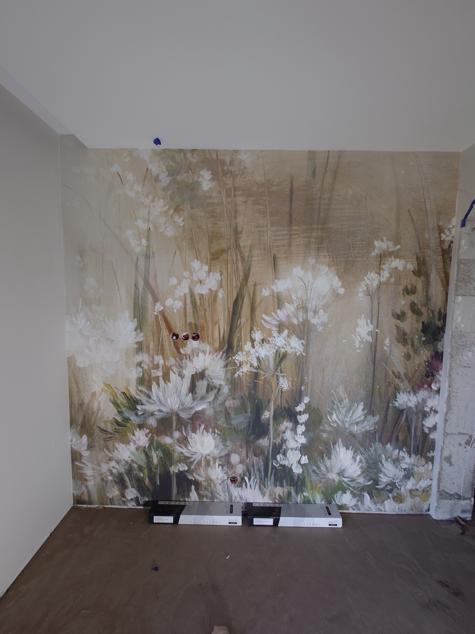 Ściana z fototapetą w kwiaty w trakcie wykańczania, widoczne puszki elektryczne i fragment niezabezpieczonej ściany. Na podłodze leżą opakowania po lampach LED.