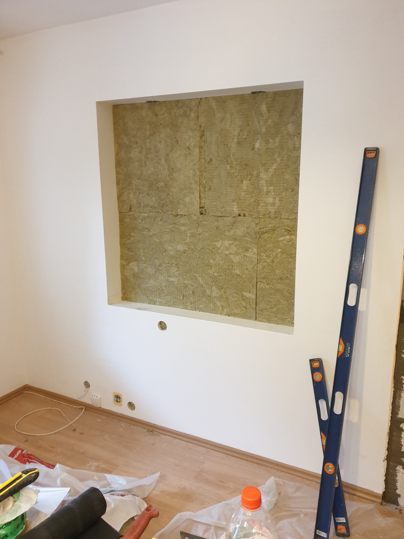 Wnęka w ścianie z izolacją z wełny mineralnej, widoczne dwa poziomy murarskie oparte o ścianę, podłoga zabezpieczona folią, gniazdka elektryczne w ścianie.