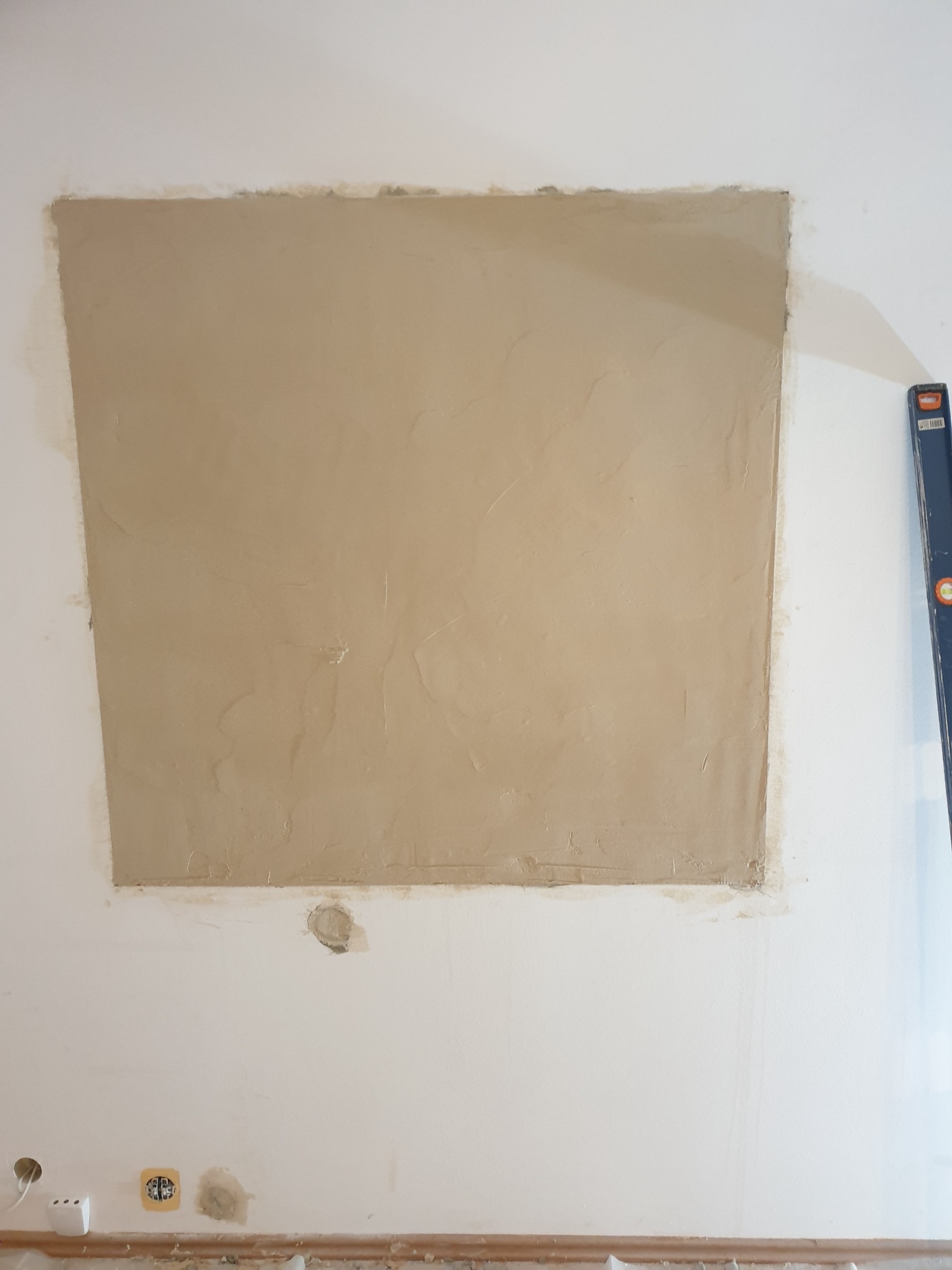Fragment ściany z nałożoną warstwą świeżej, beżowej gładzi w kształcie prostokąta; widoczne nierówności powierzchni, ślady po szpachelce i poziomica oparta o ścianę po prawej stronie