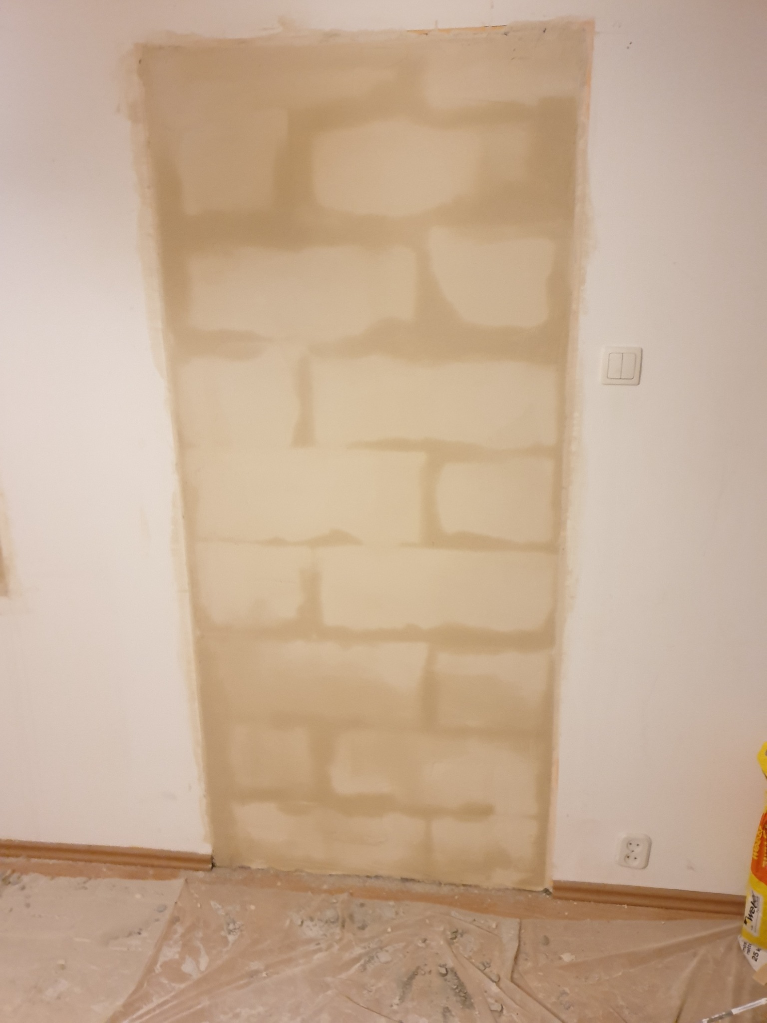 Ściana po nałożeniu pierwszej warstwy szpachli, widoczne nierówności i miejsca wymagające dodatkowej obróbki, zabezpieczona folia na podłodze.