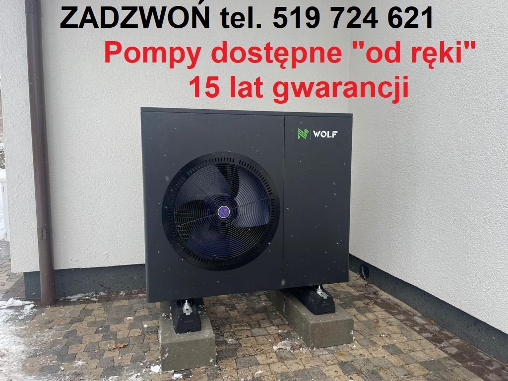 Czarna pompa ciepła marki Wolf, umieszczona na betonowych podstawach przed jasną ścianą budynku, z napisem reklamowym 'Pompy dostępne od ręki, 15 lat gwarancji' widocznym u góry.
