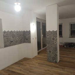 Usługi budowlano-wykończeniowe PIADA Adam Piaskowy - Częściowo wykończone wnętrze: ułożona podłoga z paneli, fragment ściany i kolumny pokryte dekoracyjnymi płytkami w geometryczne wzory, przygotowane paczki paneli podłogowych w tle.