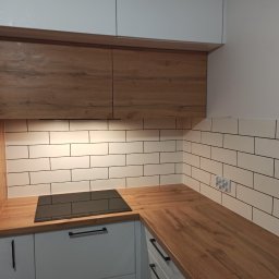 Usługi budowlano-wykończeniowe PIADA Adam Piaskowy - Nowoczesna kuchnia po remoncie: białe, podłużne płytki na ścianie, drewniany blat, czarna płyta indukcyjna, białe szafki z czarnymi uchwytami, drewniane szafki wiszące i wbudowany piekarnik...