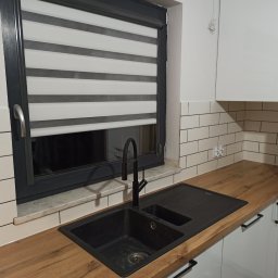 Usługi budowlano-wykończeniowe PIADA Adam Piaskowy - Czarny zlewozmywak z baterią na drewnianym blacie, białe szafki kuchenne i białe płytki imitujące cegłę, okno z roletą dzień-noc.
