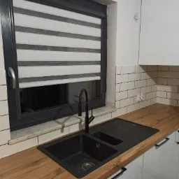 Czarny zlewozmywak z baterią na drewnianym blacie, białe szafki kuchenne i białe płytki imitujące cegłę, okno z roletą dzień-noc.