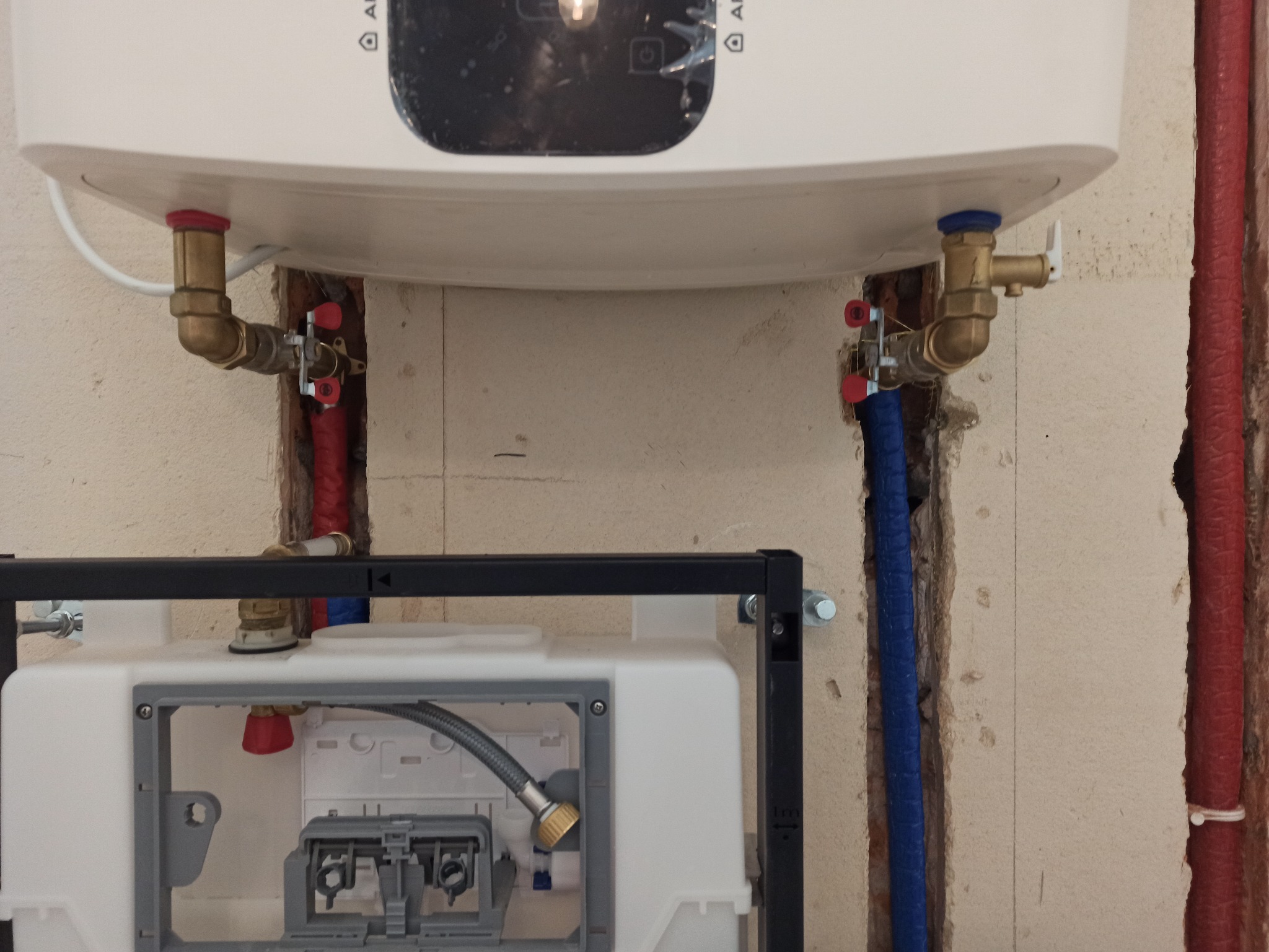 Montaż podtynkowego stelaża WC z widocznymi przyłączami wodnymi z rur PEX w kolorze niebieskim i czerwonym, zaworami kulowymi oraz bojlerem elektrycznym nad stelażem.