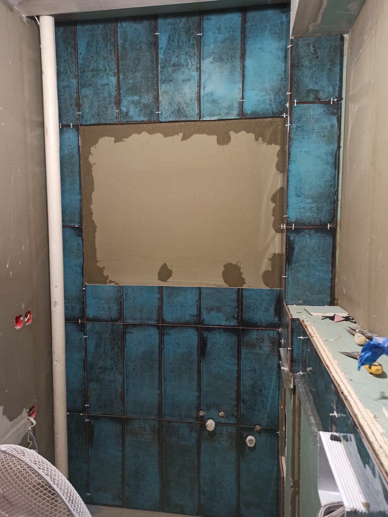 Montaż pionowych, turkusowych płytek w łazience, widoczny klej na ścianie, krzyżyki dystansowe, rury i narzędzia na blacie.