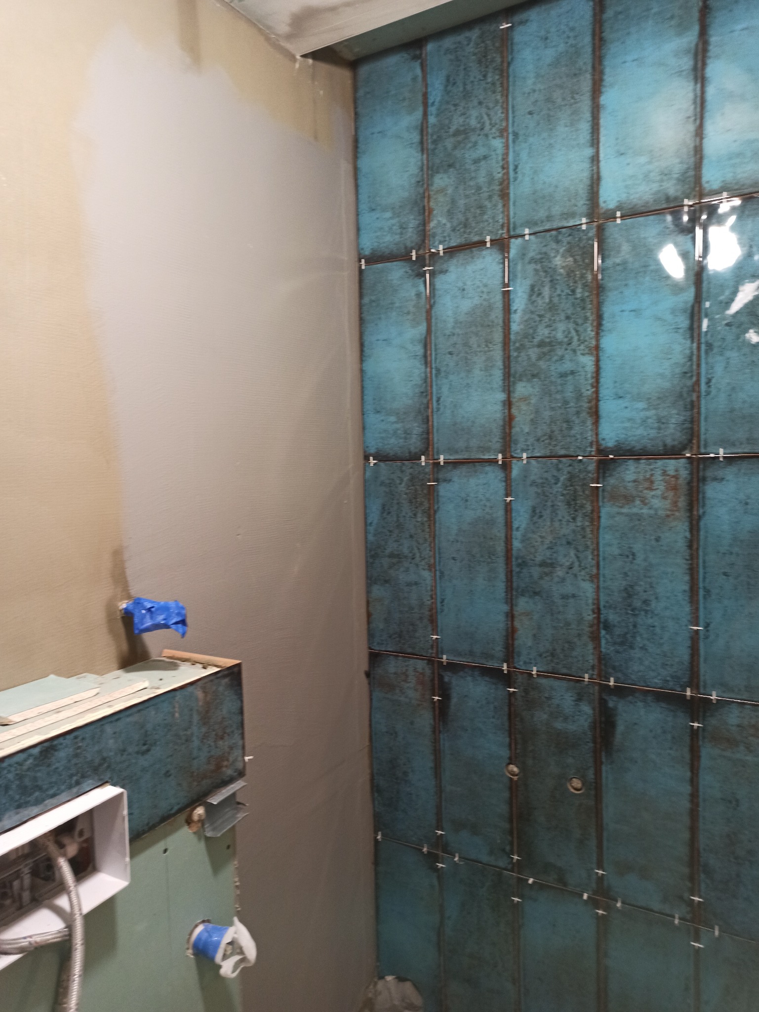 Ściana w trakcie układania płytek o metalicznym, niebiesko-brązowym wykończeniu, widoczne krzyżyki dystansowe, surowa ściana obok, przygotowania do instalacji umywalki.