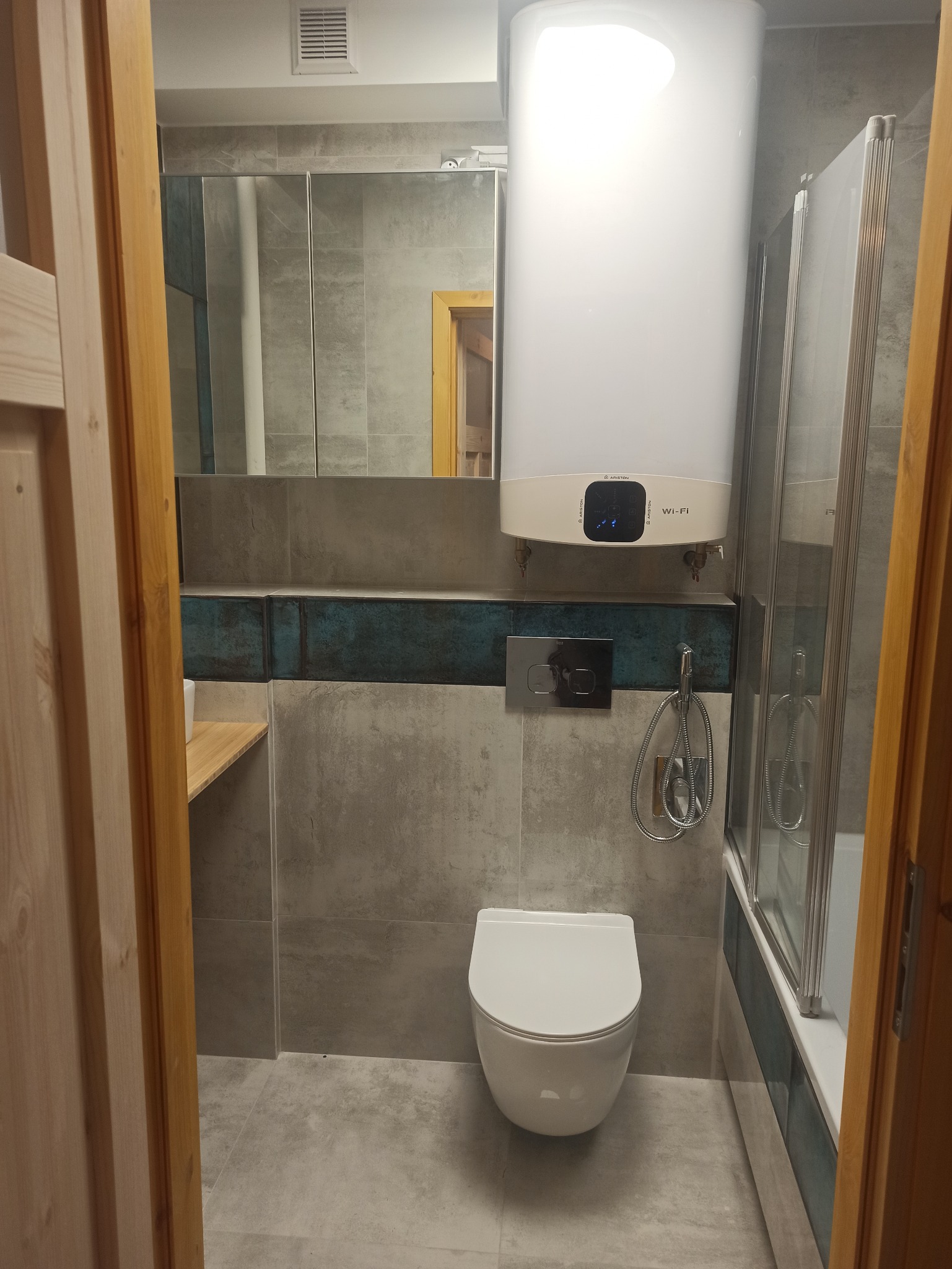 Nowoczesna łazienka z szarymi płytkami, podwieszaną toaletą i dużym bojlerem Ariston z panelem Wi-Fi, widok zza drewnianej ościeżnicy.