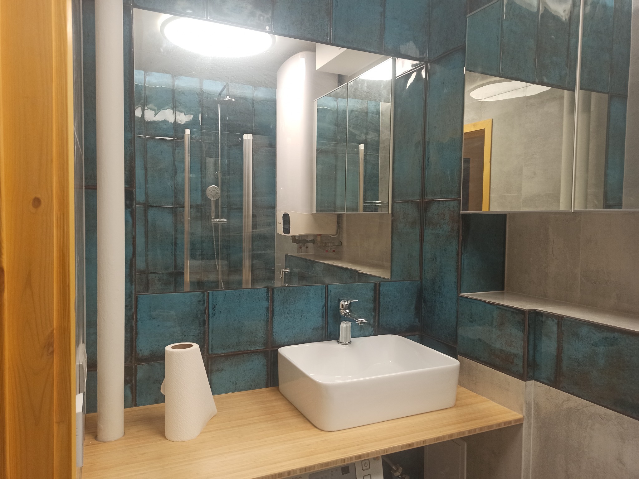 Wykończona łazienka z turkusowymi płytkami, białą umywalką nablatową i dużym lustrem z odbiciem kabiny prysznicowej oraz podgrzewacza wody.
