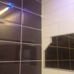 Reparexis - Fragment ściany wyłożonej błyszczącymi, ciemnymi płytkami z jasną fugą, sąsiadującej z fragmentem ściany wyłożonej jasnymi, matowymi płytkami; widoczny narożnik i fragment sufitu.