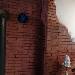 Reparexis - Ściana z imitacji cegły w odcieniach czerwieni, z fragmentem białej ściany, czarną rurą i elementami wyposażenia wnętrza, tworząca surowy, industrialny klimat.