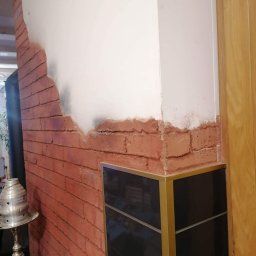 Reparexis - Fragment ściany z imitacją cegły, częściowo pokrytej gładzią, widoczny narożnik z czarną szafką i złotymi profilami oraz fragment drewnianych drzwi.