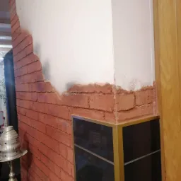 Fragment ściany z imitacją cegły, częściowo pokrytej gładzią, widoczny narożnik z czarną szafką i złotymi profilami oraz fragment drewnianych drzwi.
