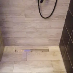 Wykończona strefa prysznicowa z odpływem liniowym i baterią natryskową w kolorze czarnym, płytki imitujące drewno, widok z góry.