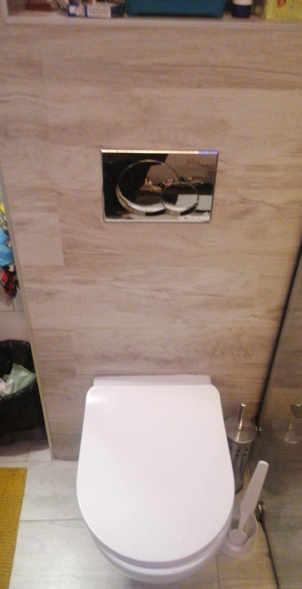 Wisząca toaleta z chromowanym przyciskiem spłukiwania na ścianie imitującej drewno, widoczny fragment dywanika i szczotka do WC