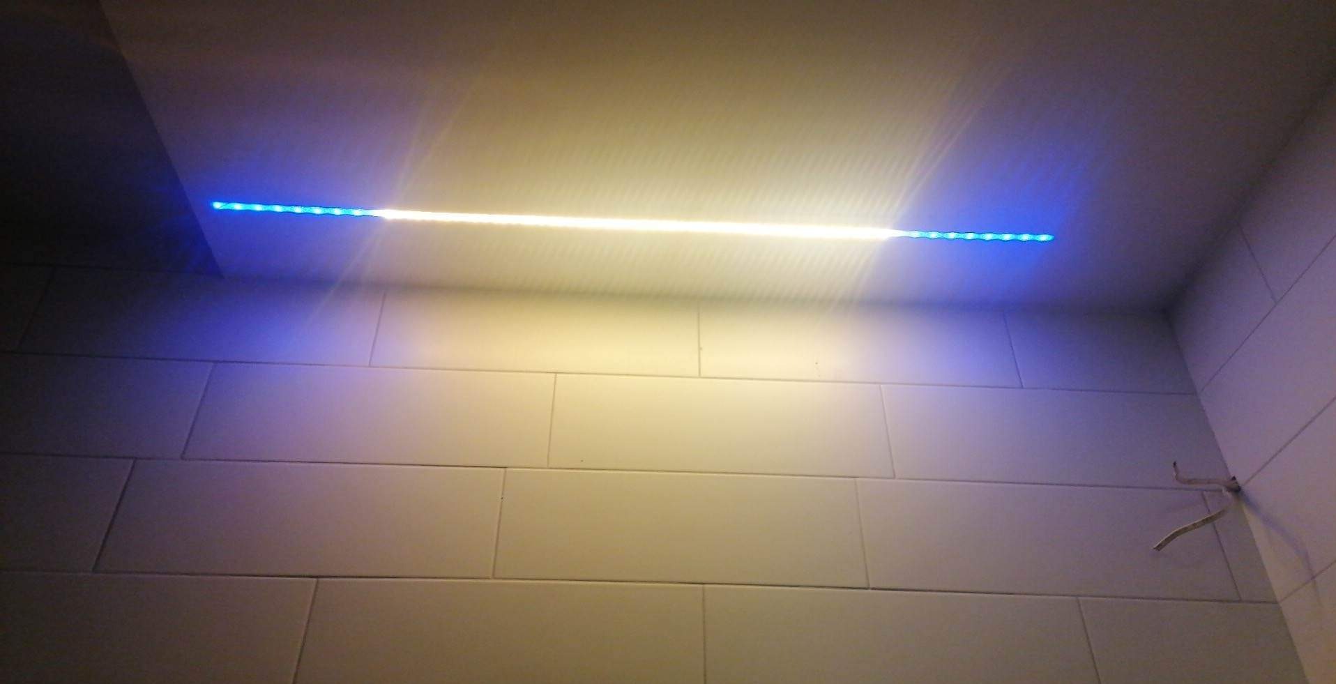 Świecąca listwa LED, biało-niebieska, zamontowana pod sufitem w łazience wyłożonej białymi, prostokątnymi płytkami; widoczne wystające przewody elektryczne.