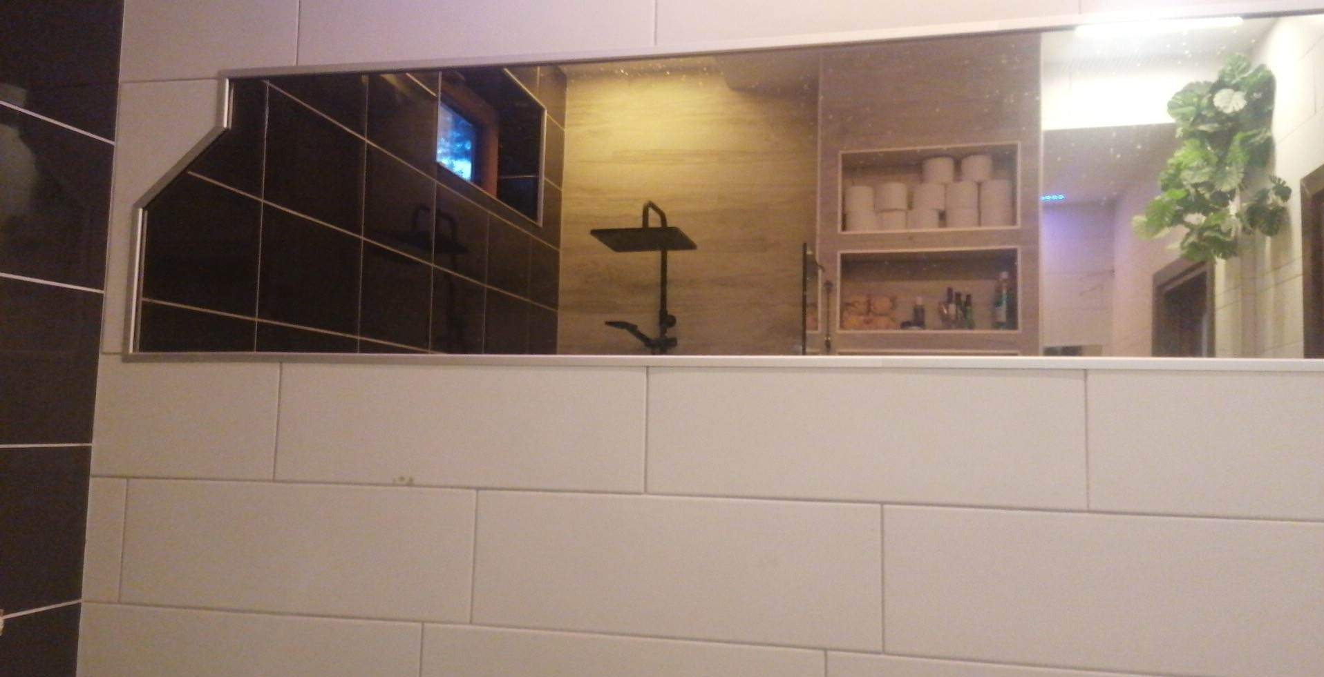 Białe, prostokątne płytki ścienne z widocznym odbiciem w lustrze: czarna kabina prysznicowa, półka z zapasami i dekoracyjna roślina.