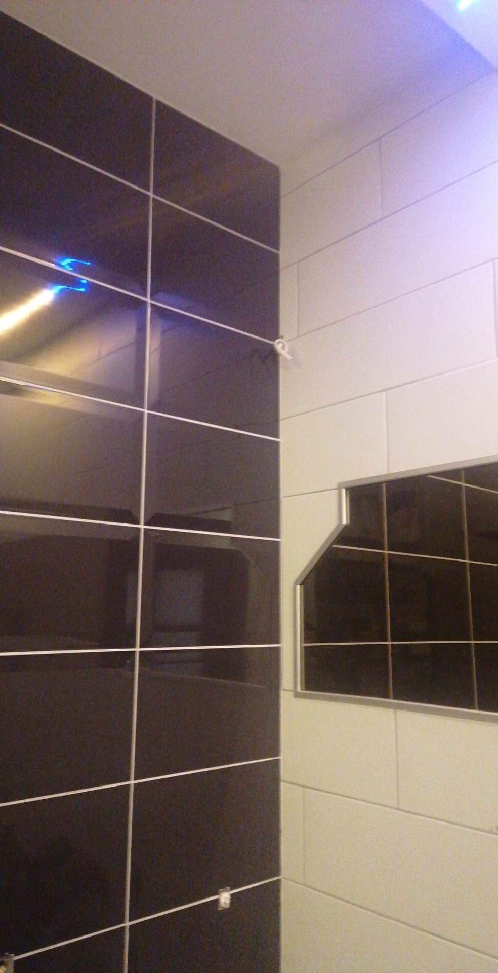 Fragment ściany wyłożonej błyszczącymi, ciemnymi płytkami z jasną fugą, sąsiadującej z fragmentem ściany wyłożonej jasnymi, matowymi płytkami; widoczny narożnik i fragment sufitu.