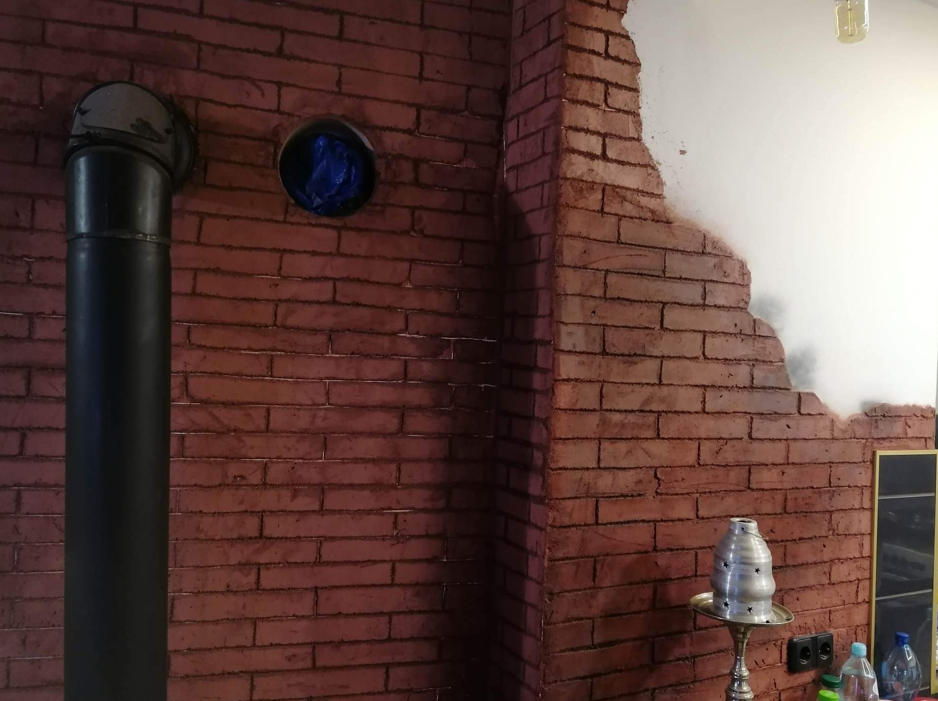 Ściana z imitacji cegły w odcieniach czerwieni, z fragmentem białej ściany, czarną rurą i elementami wyposażenia wnętrza, tworząca surowy, industrialny klimat.