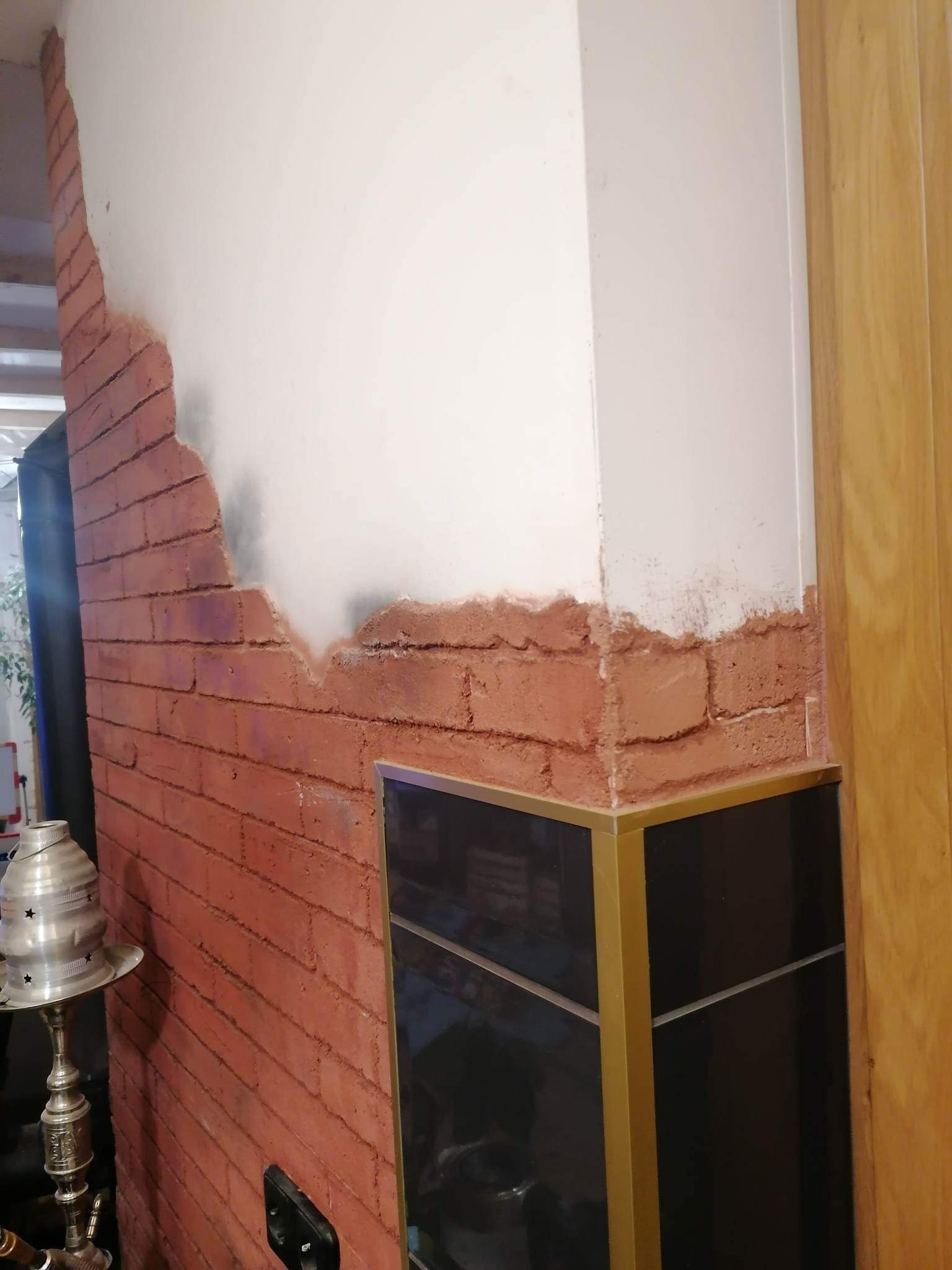 Fragment ściany z imitacją cegły, częściowo pokrytej gładzią, widoczny narożnik z czarną szafką i złotymi profilami oraz fragment drewnianych drzwi.