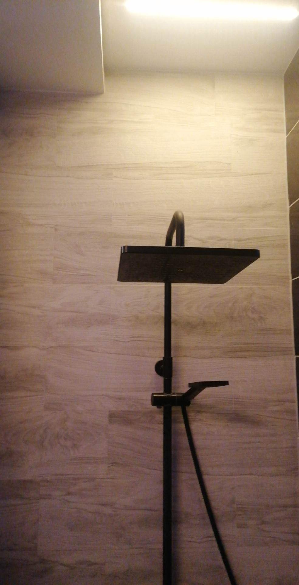 Czarna, kwadratowa deszczownica zamontowana na ścianie wyłożonej płytkami imitującymi drewno, oświetlona górnym światłem.