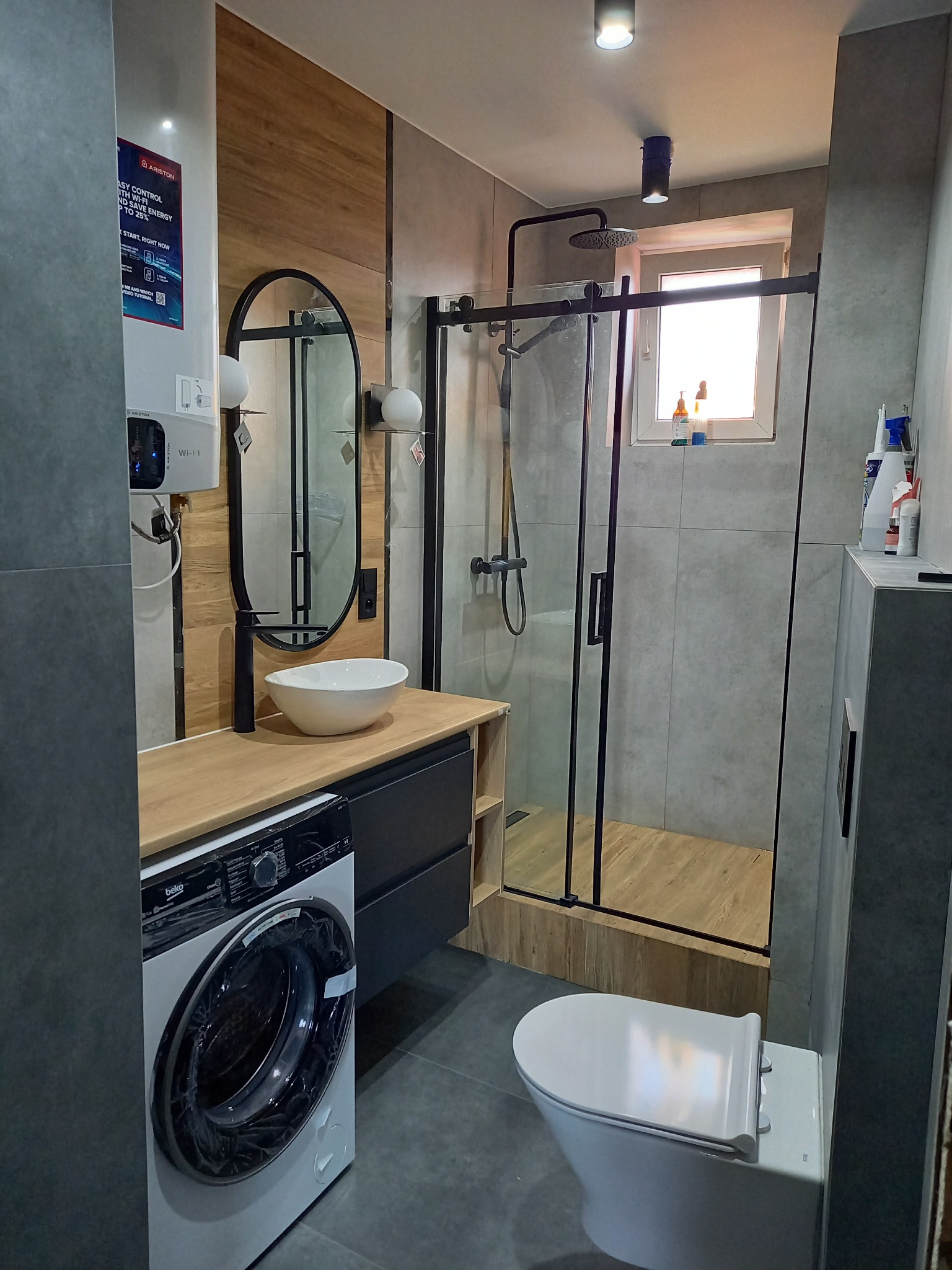 Nowoczesna łazienka po remoncie: kabina prysznicowa z czarnymi ramami, umywalka nablatowa na drewnianym blacie, toaleta wisząca i pralka Beko z folią zabezpieczającą na drzwiczkach.