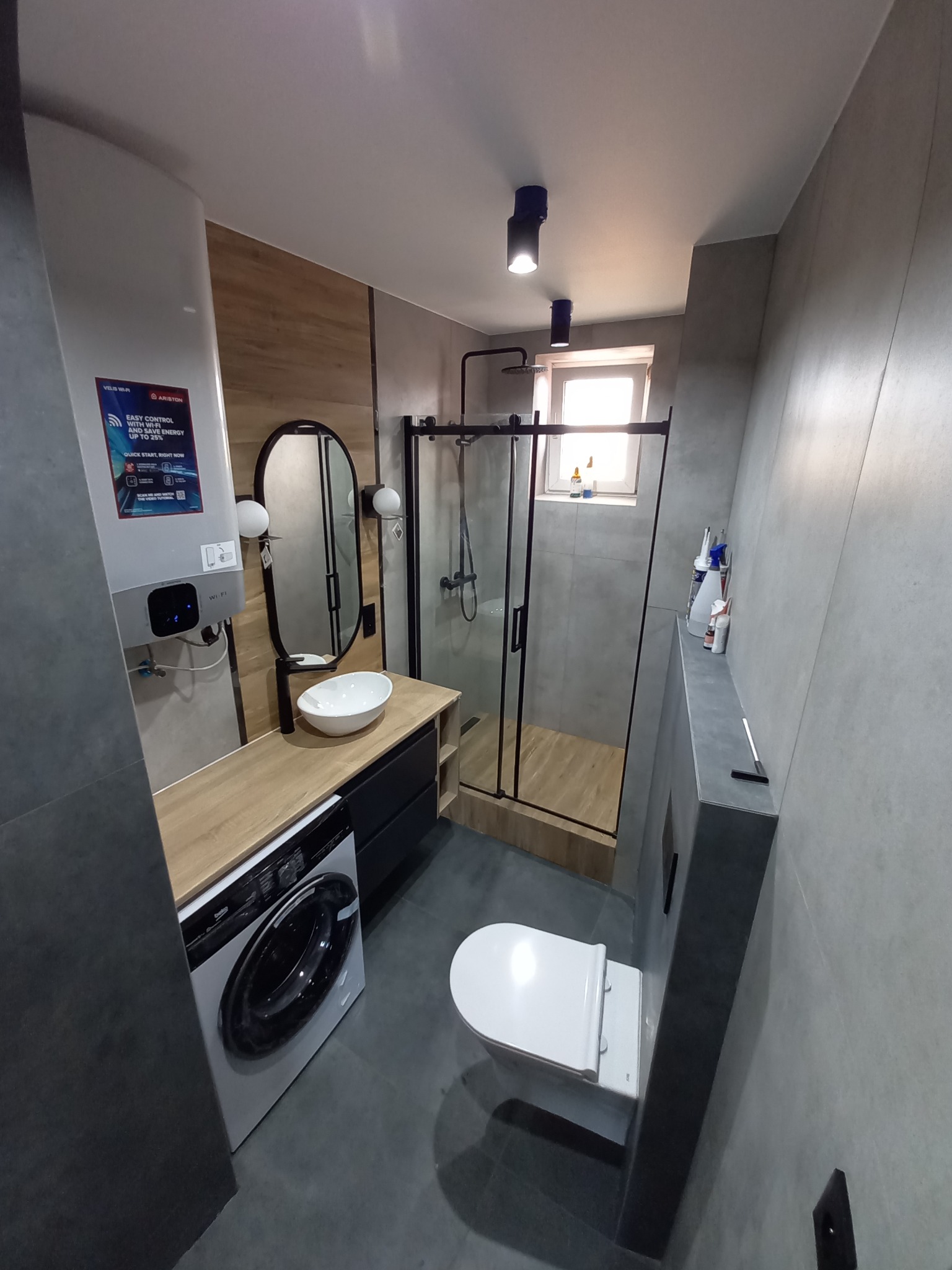Nowoczesna łazienka z szarą płytką, kabiną prysznicową z czarnymi ramami, umywalką nablatową na drewnianym blacie, pralką i wiszącą toaletą, oświetlona punktowymi lampami sufitowymi.
