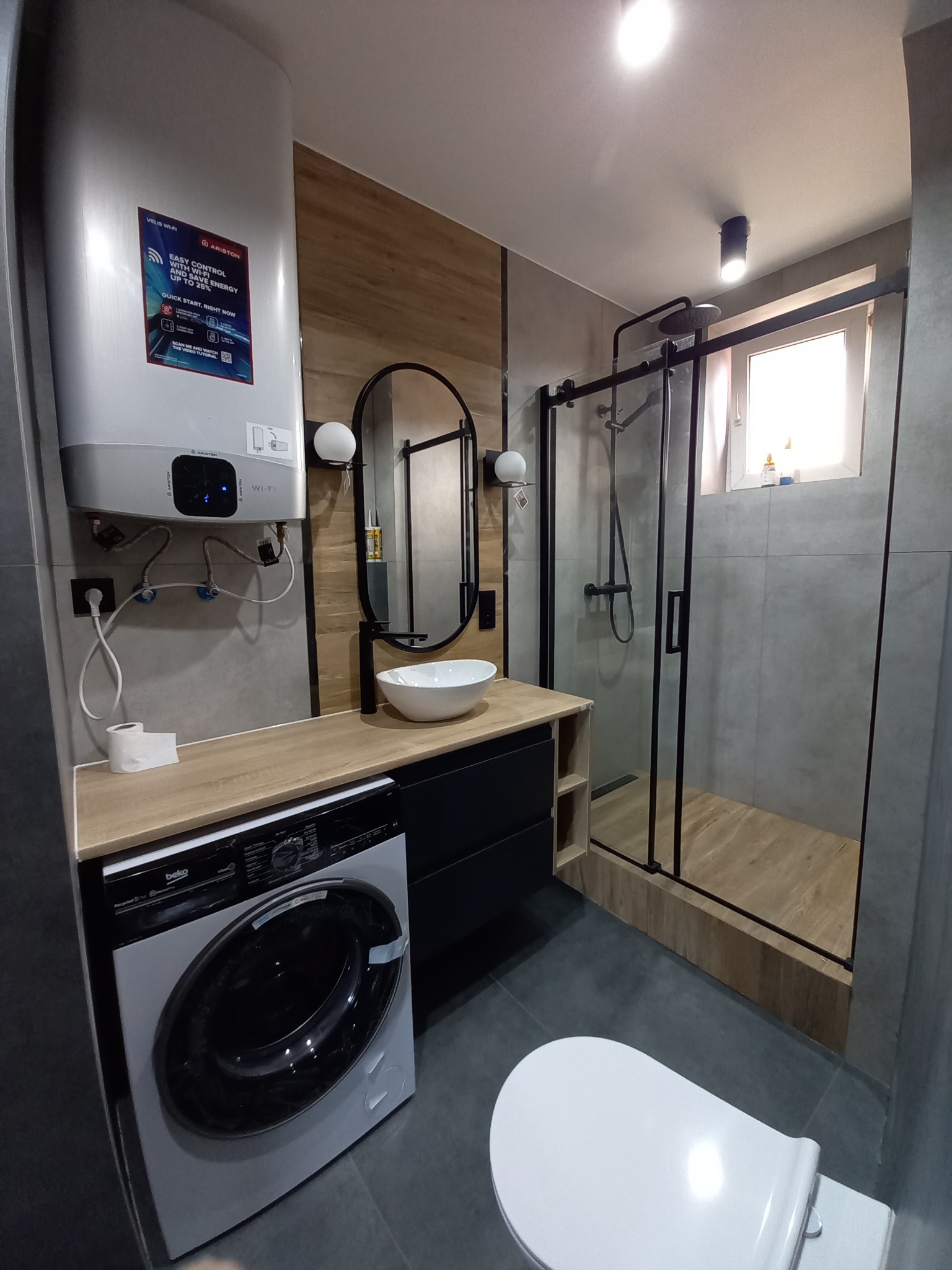 Nowoczesna łazienka z szarymi płytkami, drewnopodobną zabudową, kabiną prysznicową z czarnymi ramami, wiszącą umywalką, pralką i podgrzewaczem wody Ariston Velis Wi-Fi.