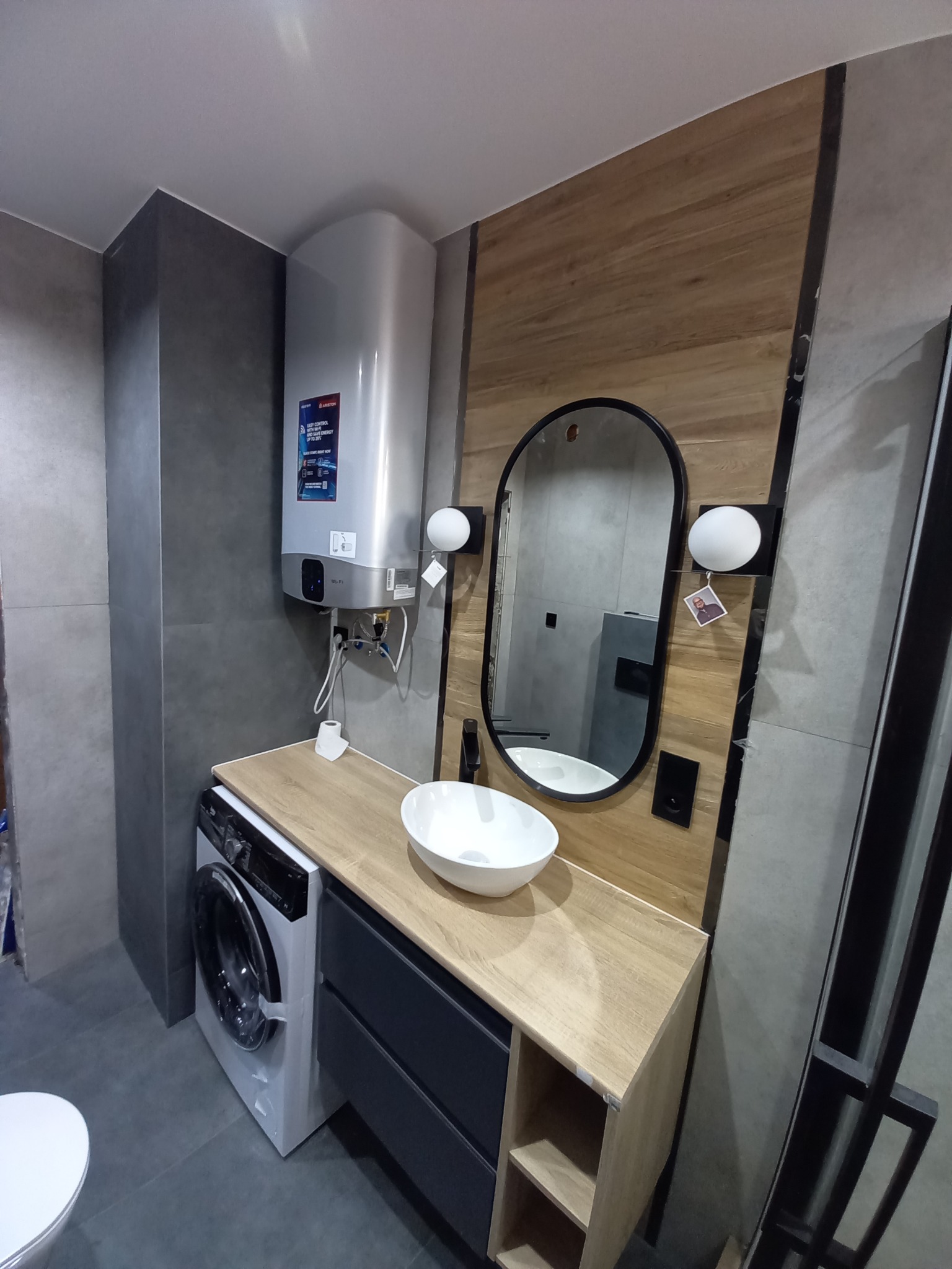 Nowoczesna łazienka z drewnianymi akcentami: umywalka nablatowa, lustro w czarnej ramie, pralka pod blatem i bojler Ariston na ścianie.