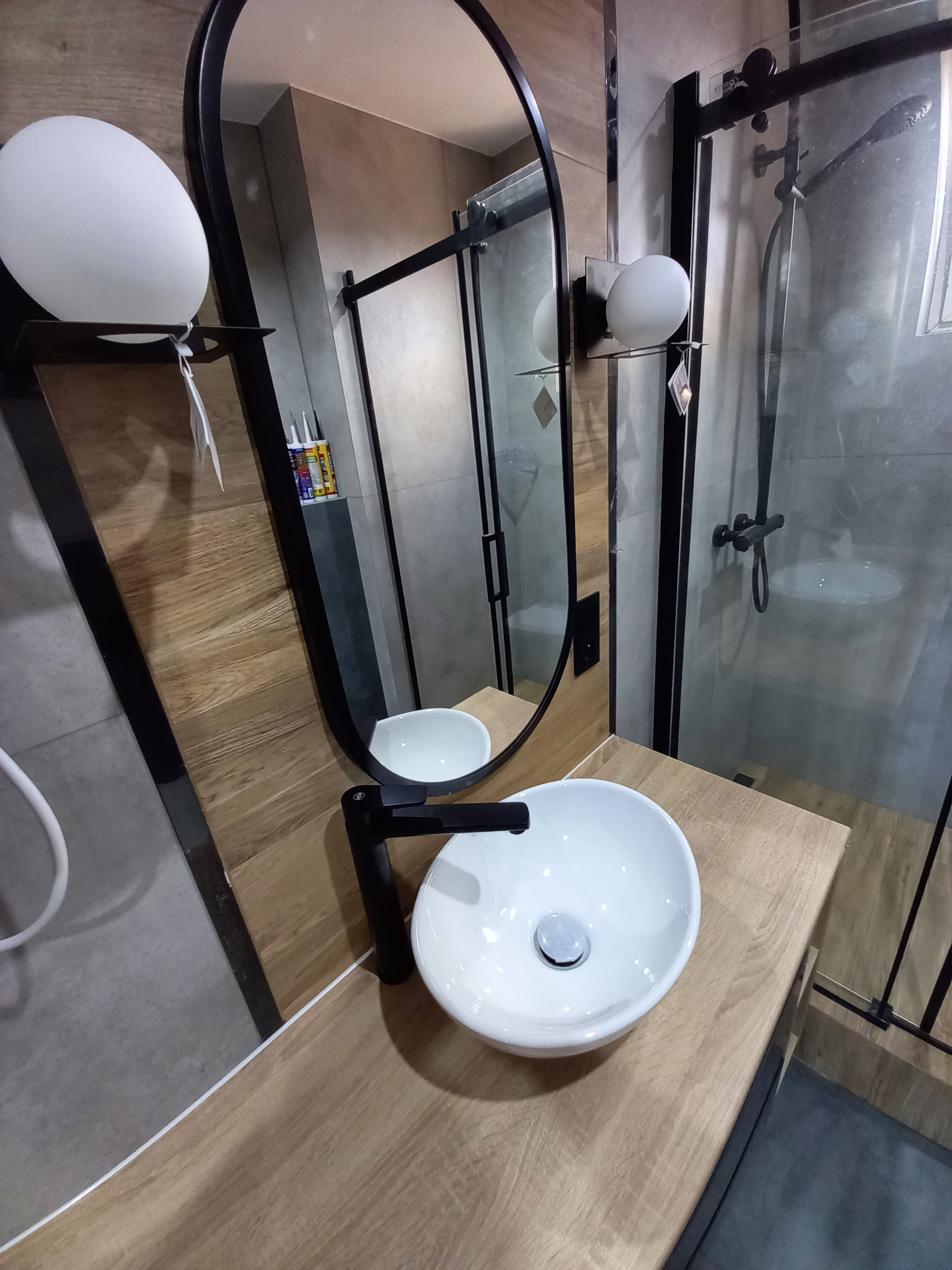 Nowoczesna łazienka z owalną białą umywalką nablatową, czarną armaturą i kabiną prysznicową z czarnymi profilami, drewnopodobne panele ścienne i blat.