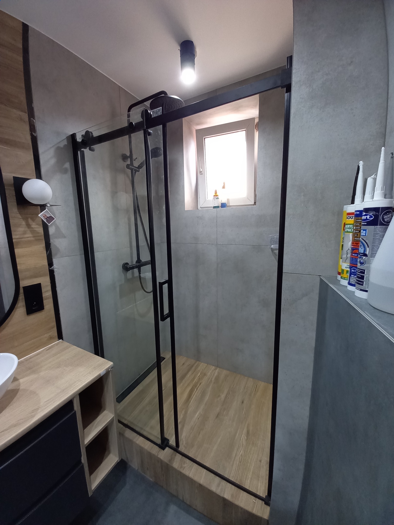 Nowoczesna kabina prysznicowa z czarnymi ramami i drewnopodobną podłogą, widoczne silikony i uszczelniacze na półce, szare płytki na ścianach, minimalistyczna umywalka z szafką w tle.