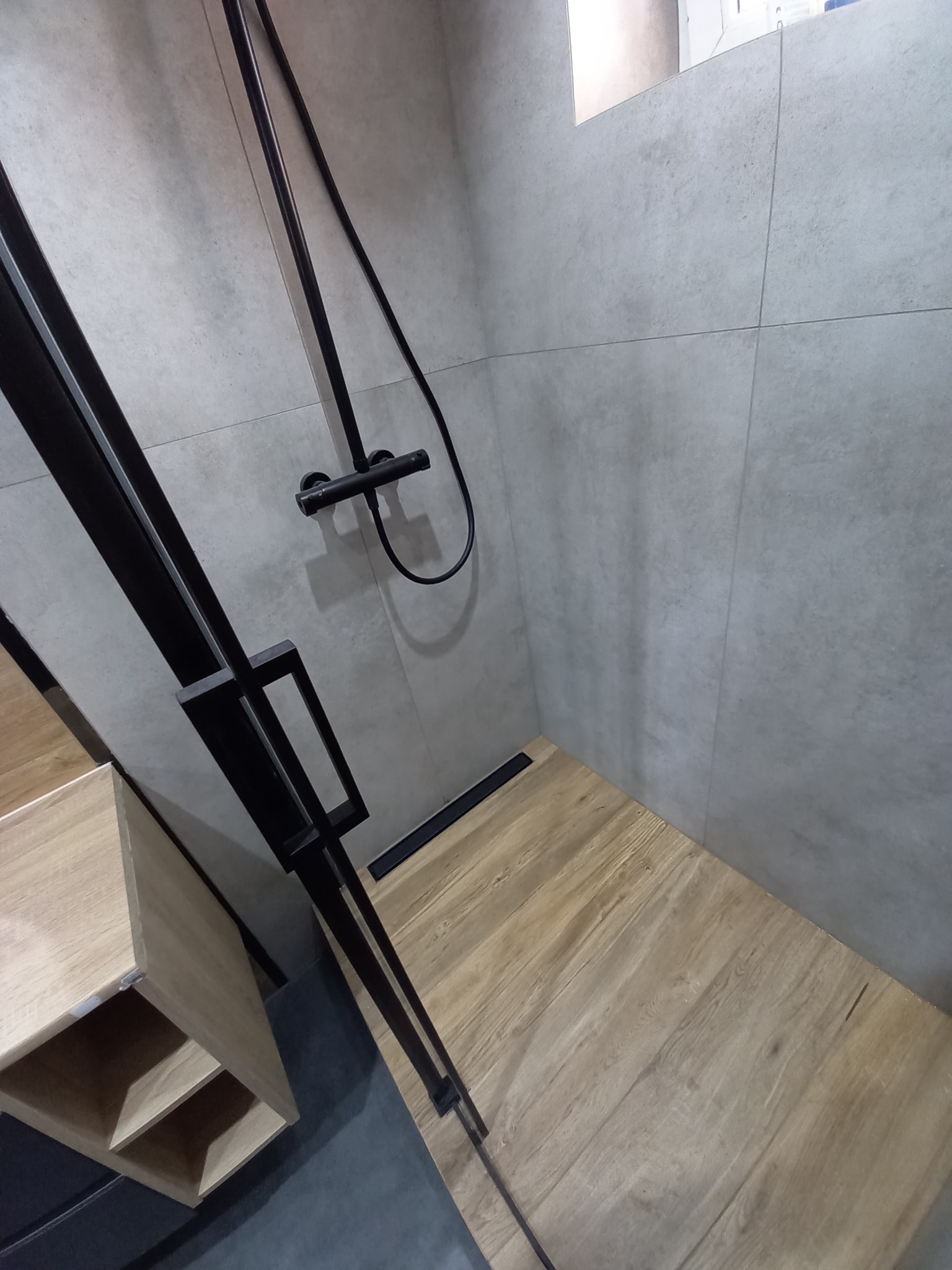 Nowoczesna kabina prysznicowa z czarną armaturą i odpływem liniowym, wykończona płytkami imitującymi drewno i szarą ceramiką.