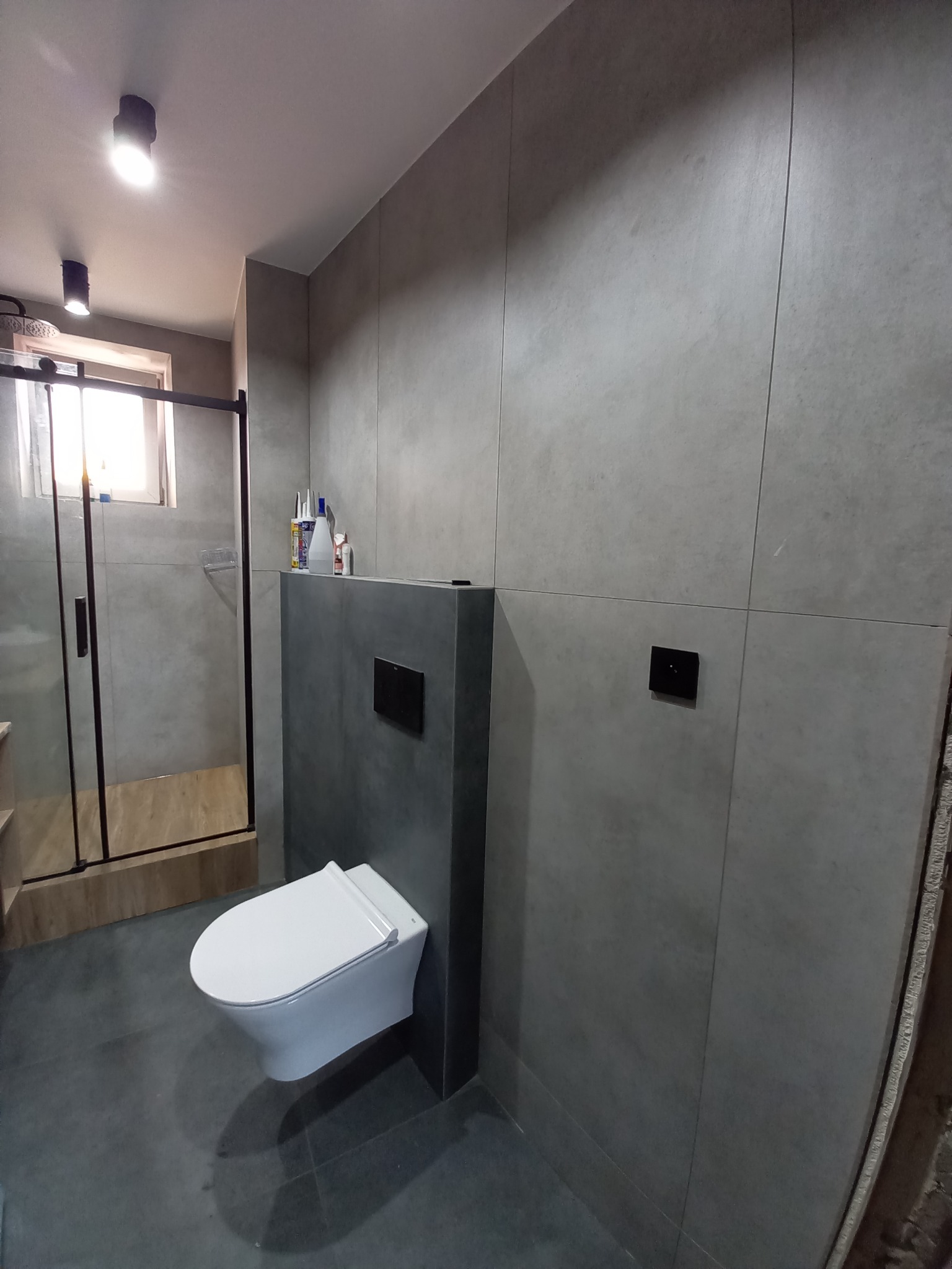 Nowoczesna łazienka z szarymi płytkami, wiszącą toaletą i kabiną prysznicową z czarnymi ramami, oświetlona punktowymi lampami sufitowymi.