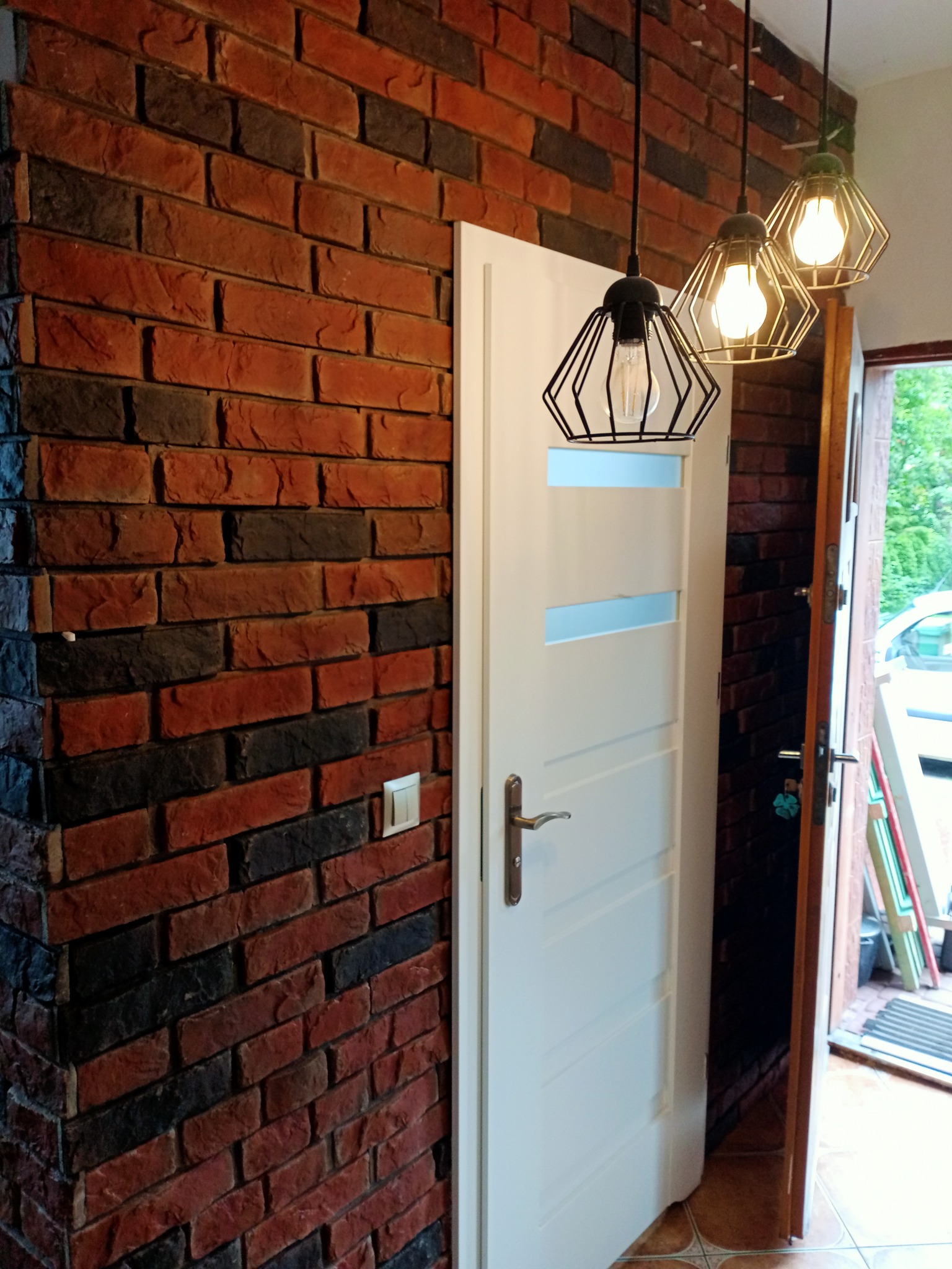 Ściana z cegły klinkierowej w odcieniach czerwieni i czerni, sąsiadująca z białą, częściowo otwartą, nowoczesną stolarką drzwiową z przeszkleniami i wiszącymi lampami.