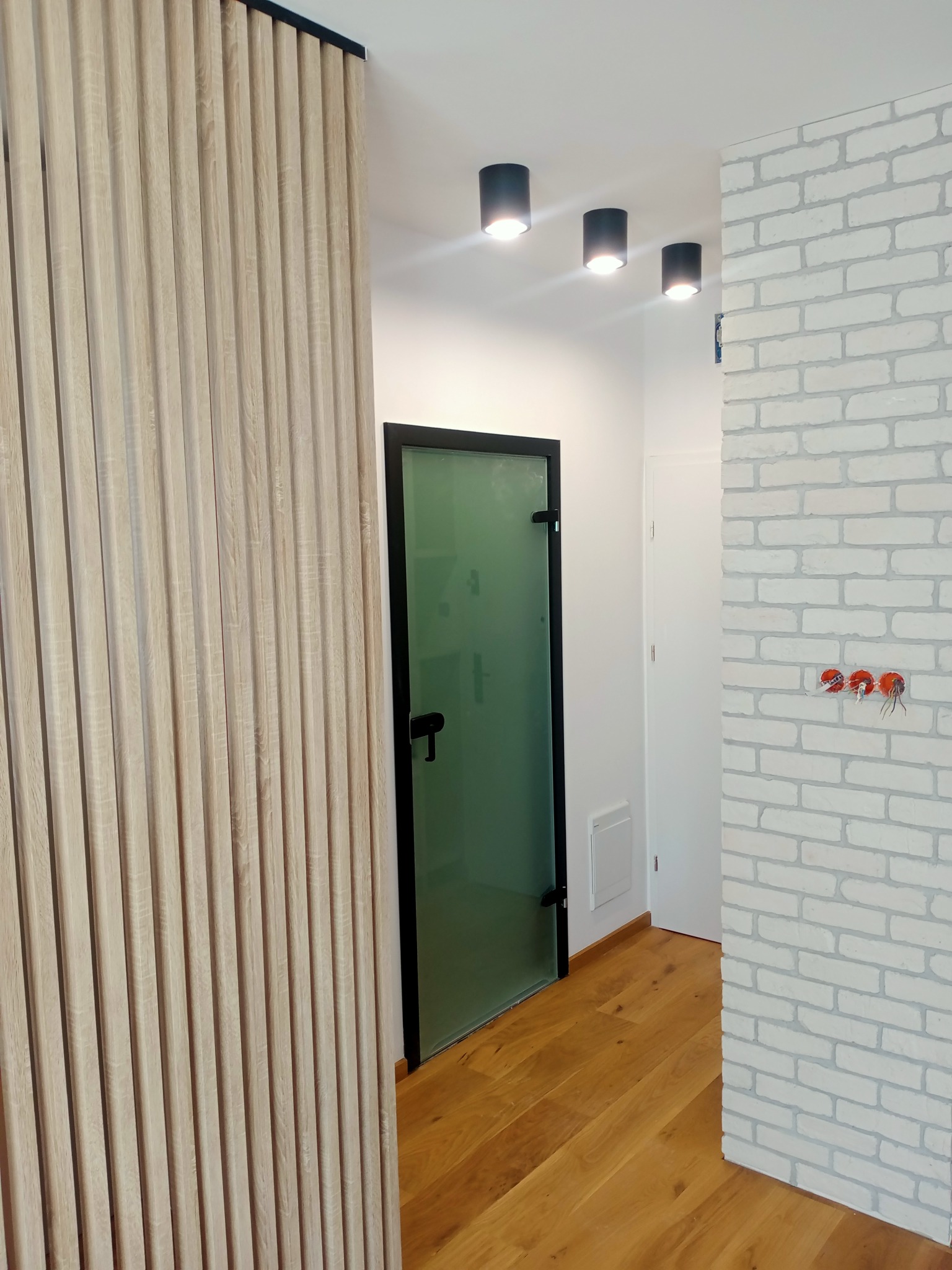 Nowoczesne wnętrze: drewniane lamele, szklane drzwi w czarnej ramie, biała cegła i drewniana podłoga. Widoczne czarne lampy sufitowe i gniazdka elektryczne.