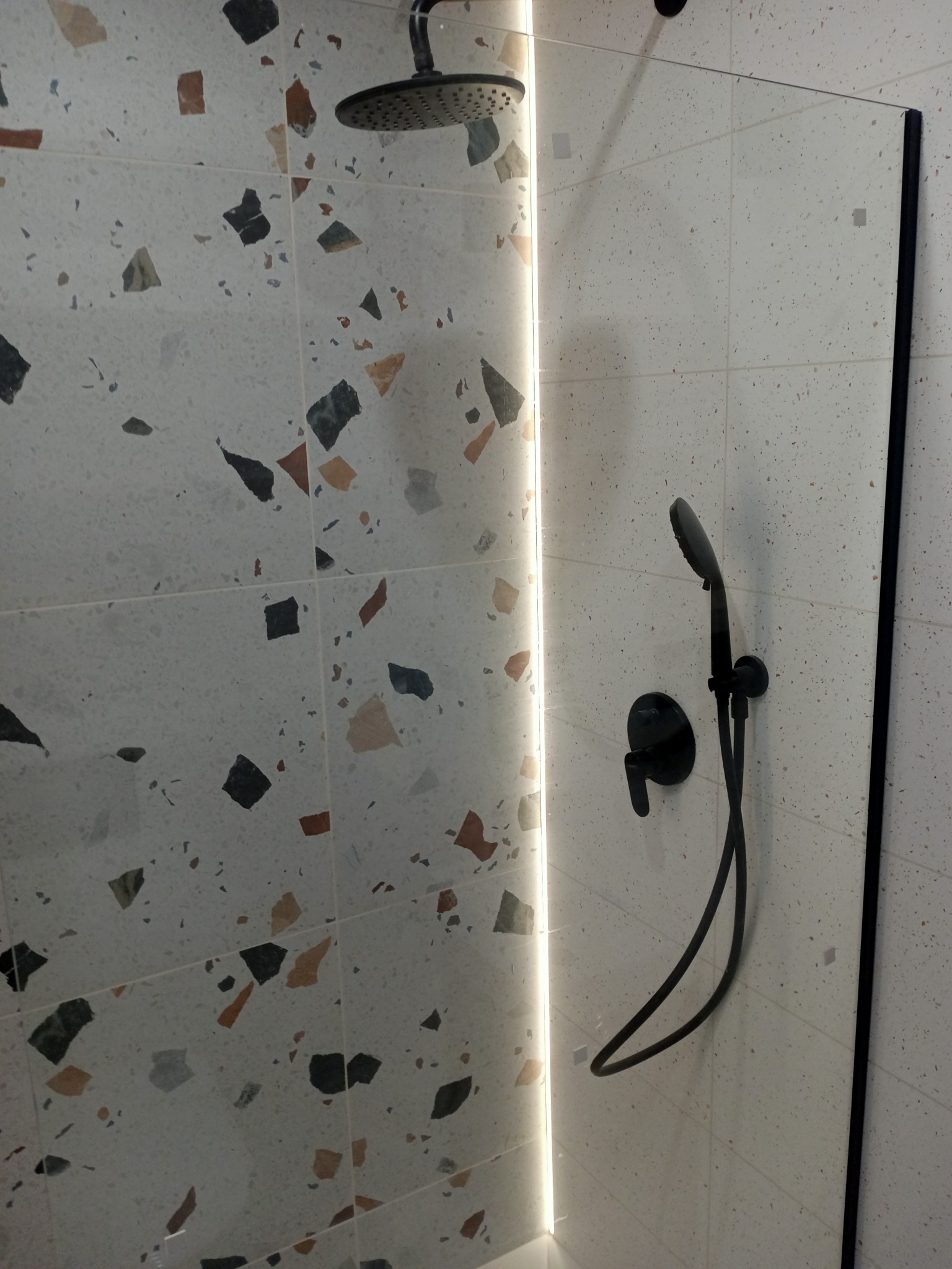 Wykończona kabina prysznicowa z czarną armaturą na tle płytek lastryko i podświetlaną listwą LED. Nowoczesny design łazienki.