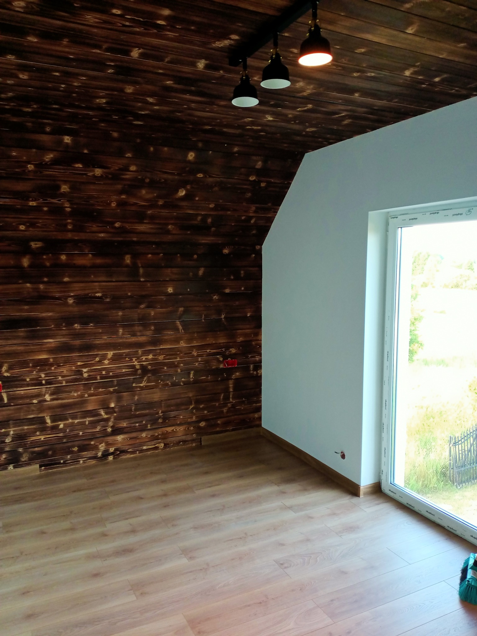 Wykończone wnętrze poddasza z drewnianą boazerią na ścianie i suficie, jasnymi panelami podłogowymi i oknem PCV. Widoczne lampy sufitowe i gładka, biała ściana.