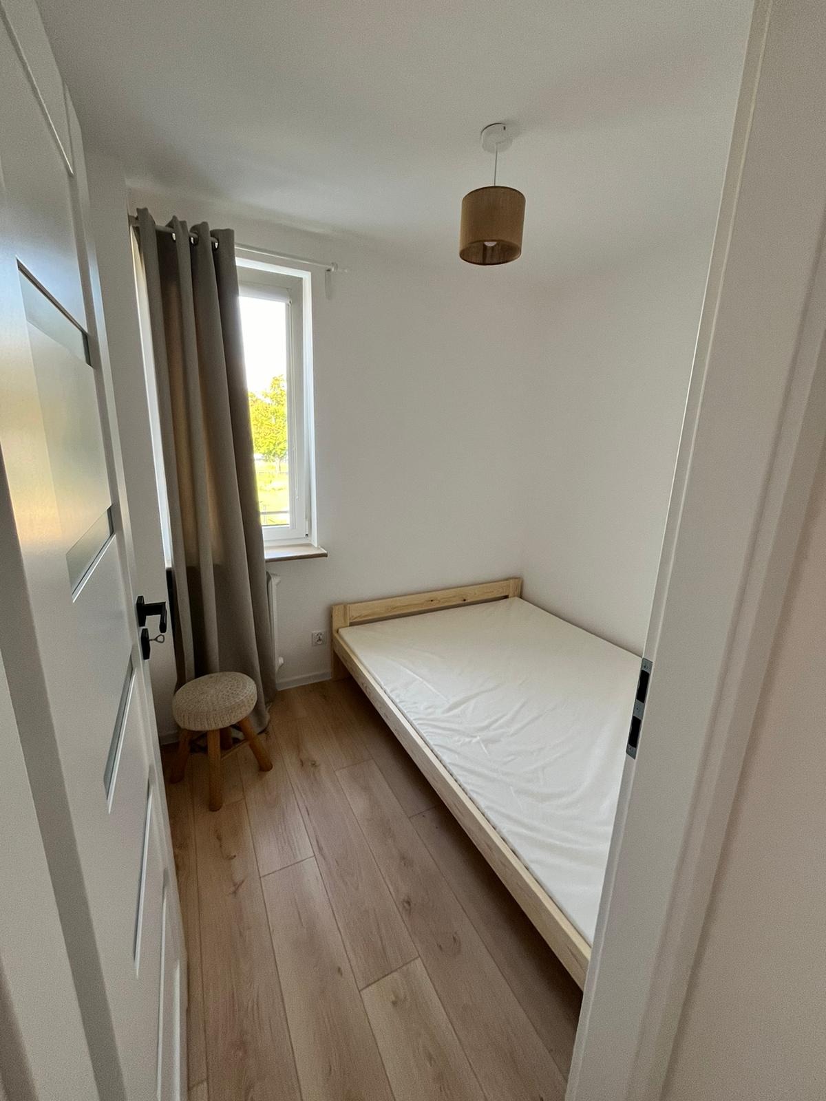 Wnętrze pokoju po remoncie: drewniana podłoga, łóżko z materacem, okno z zasłonami i lampa. Widok zza uchylonych drzwi, ukazujący schludne wykończenie.