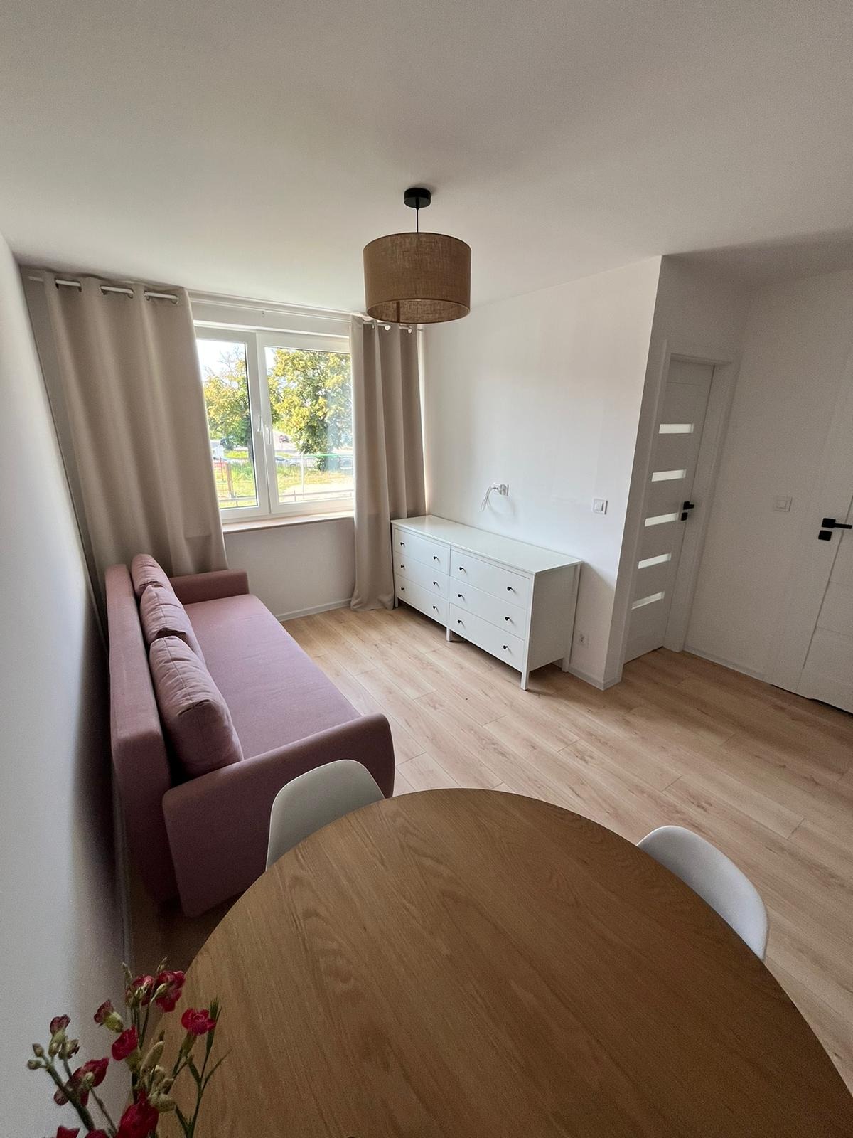 Jasne wnętrze po remoncie: różowa sofa, drewniany stół, biała komoda i panele podłogowe. Widok na okno z beżowymi zasłonami i lampa w stylu boho.